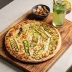 A professional top-down photograph of a grilled zucchini pizza with red sauce on a rustic wooden board, served with a side of mixed pickles and a refreshing glass of mint lemonade. Healthy vegan food option from Vegans Hub.