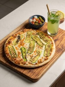 A professional top-down photograph of a grilled zucchini pizza with red sauce on a rustic wooden board, served with a side of mixed pickles and a refreshing glass of mint lemonade. Healthy vegan food option from Vegans Hub.